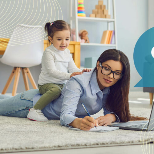 A woman writes on a laptop in front of an open laptop on a rug, while her child sits on her back