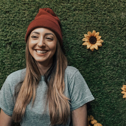A woman wearing a red hat and gray T-shirt stands against a wall with greenery and yellow sunflowers