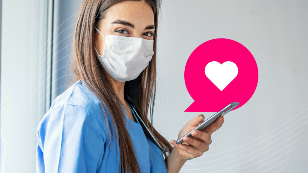 A nurse with long brown hair and blue scrubs and a white mask holding a smartphone