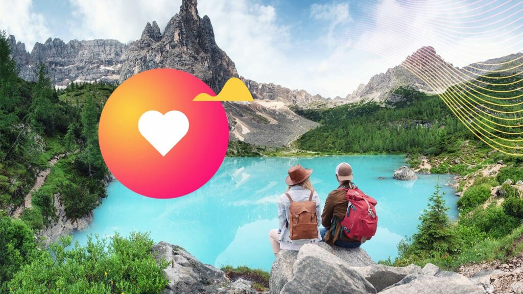 A couple wearing backpacks sit on a rock overlooking a turquoise colored lake and mountains