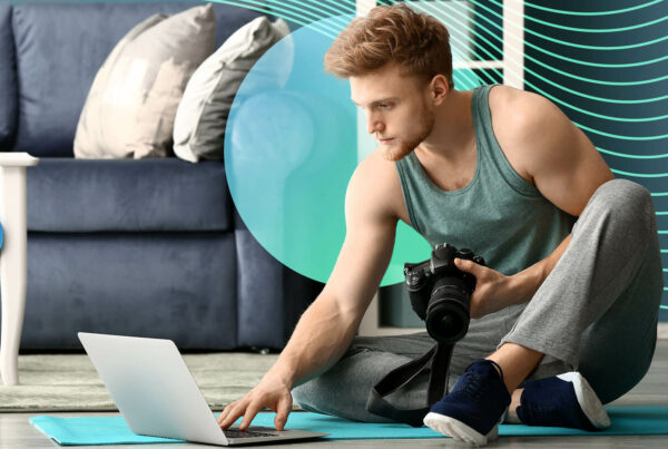 A male fitness influencer types on a laptop while sitting on an exercise mat on the floor next to a sofa