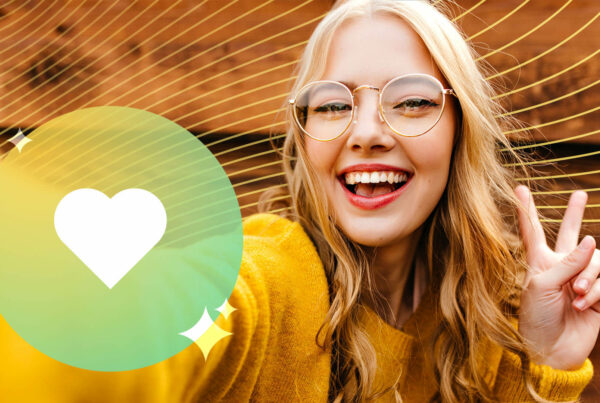A blonde woman with a yellow sweater holds up a peace sign and smiles for a selfie; green circle with white heart icon