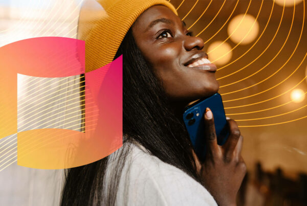A Black woman wearing a yellow beanie and white top holds her phone below her chin and smiles