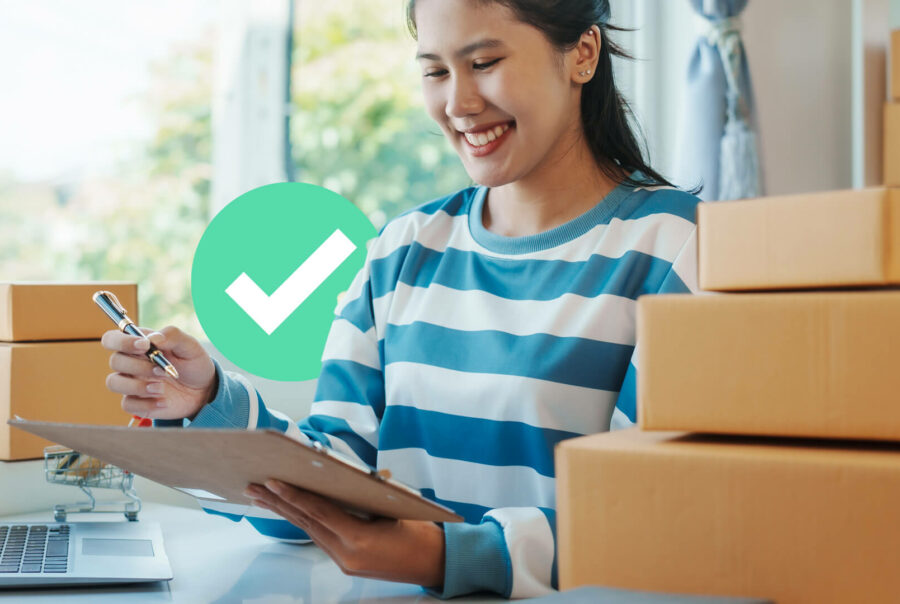A woman checks off an item on a clipboard while standing next to shipping boxes and a laptop
