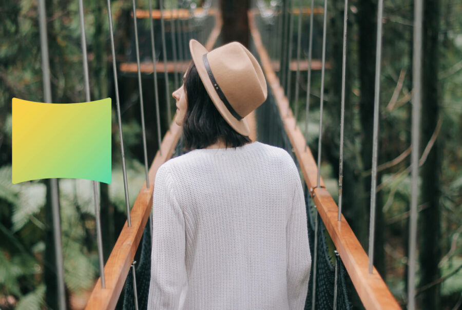 A woman in a white top and a hat walks over a bridge in a forest